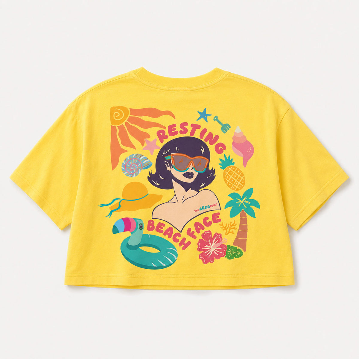 A bright yellow short-sleeve crop top t-shirt featuring a colorful retro-style graphic of a woman with sunglasses surrounded by beach-themed illustrations like a palm tree, pineapple, and pool float, with the text 'RESTING BEACH FACE'.